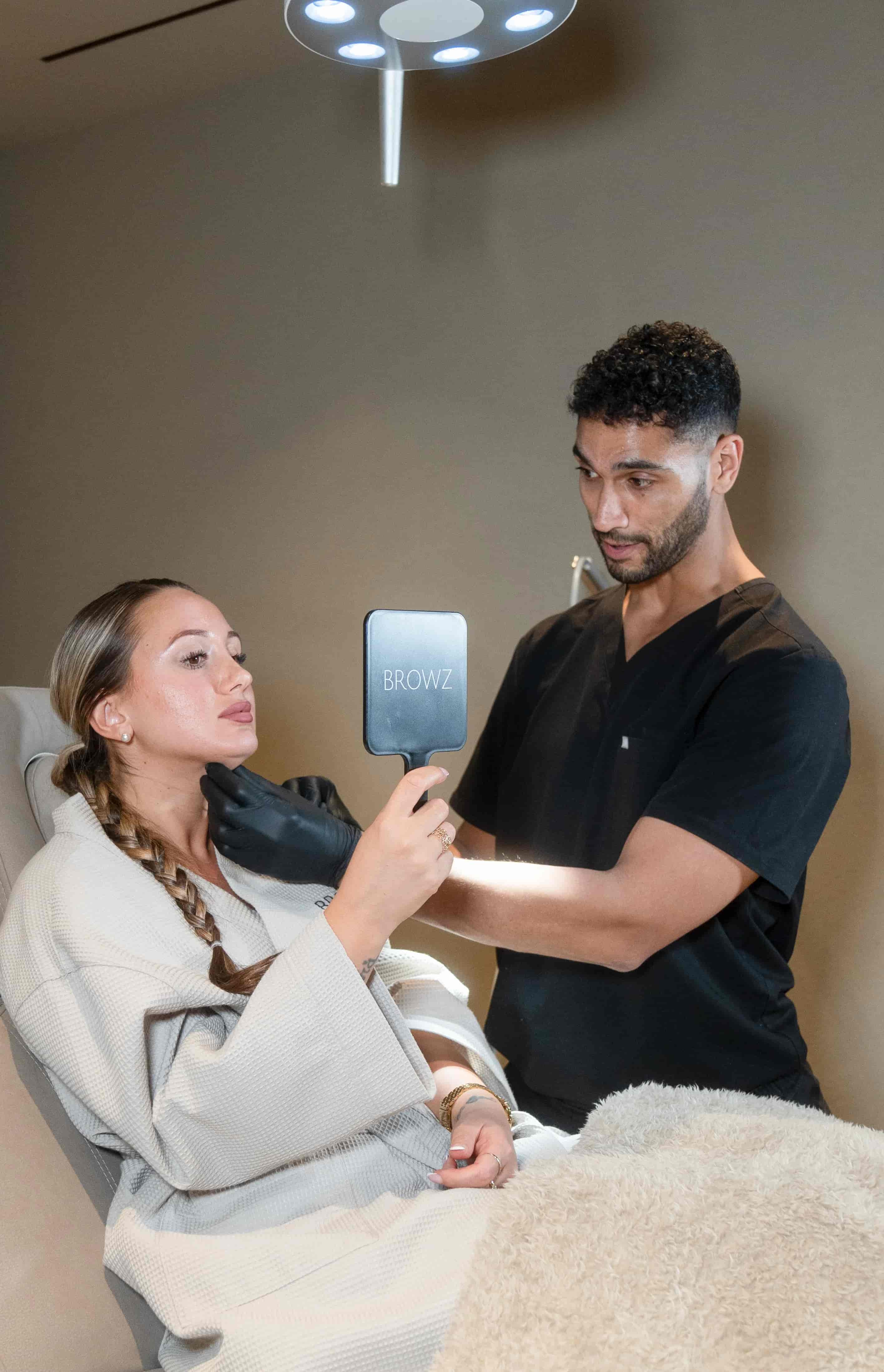 A professional performing a Sculptra treatment on a client, with the client holding a mirror to check results at BROWZ in Dubai