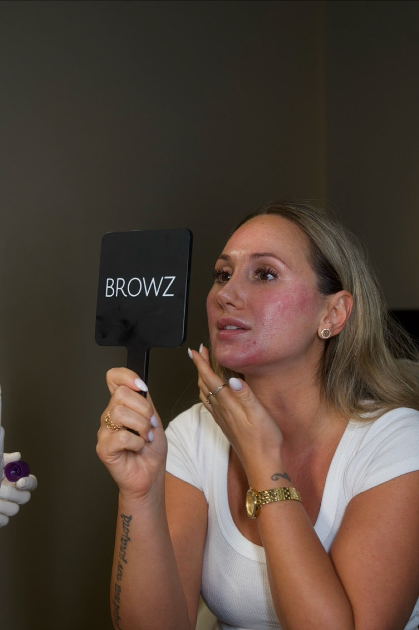 A female client amazed by the instant effects of Radiesse filler to her face after treatment while holding a mirror.
