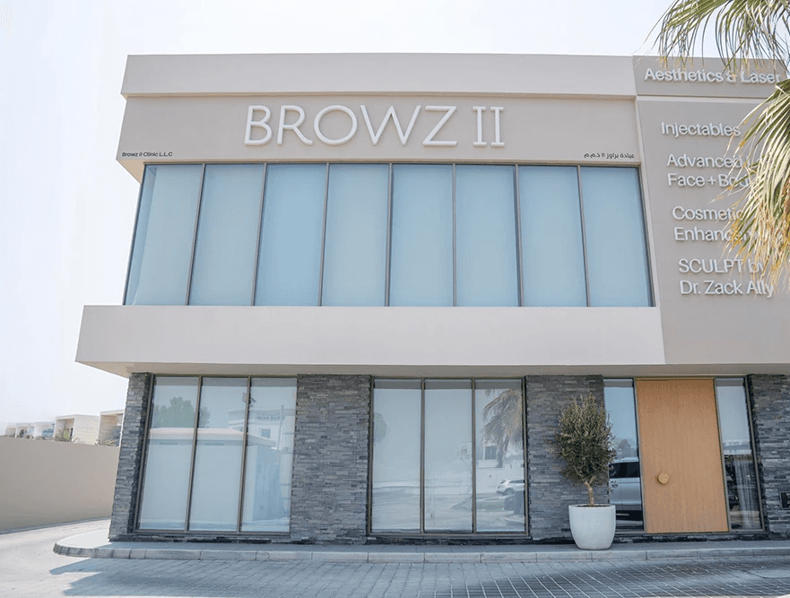 BROWZ II aesthetic clinic exterior featuring a modern beige facade, large light blue-tinted windows, gray stone-textured lower wall panels, a light wood entrance door, and a potted palm on a paved forecourt.