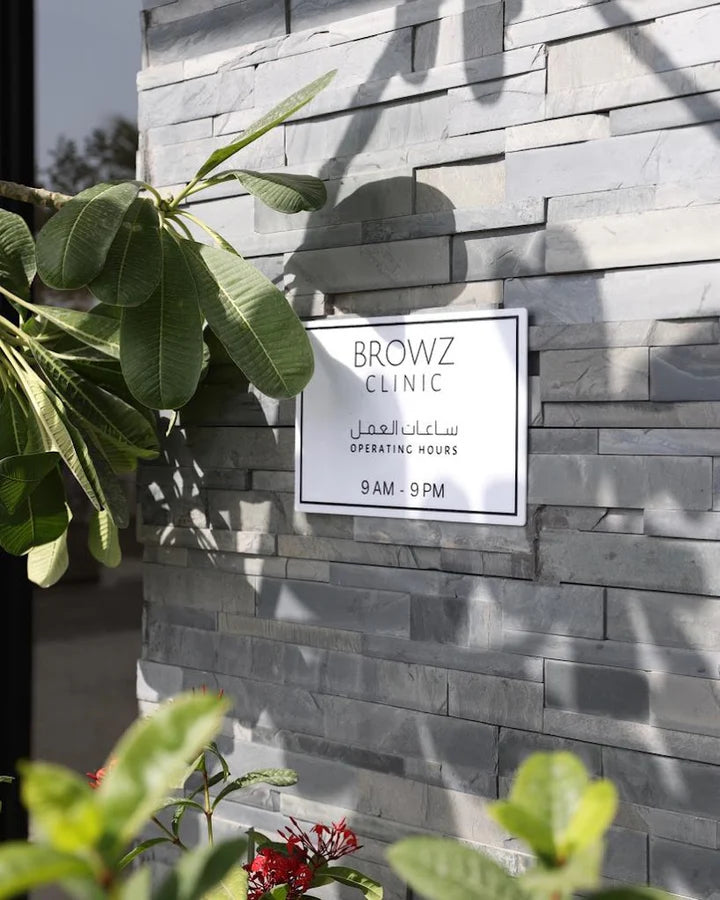 A white rectangular sign reading 'BROWZ CLINIC' with Arabic text and 'OPERATING HOURS 9 AM - 9 PM' mounted on a gray stacked-stone wall, with green leaves in the foreground.